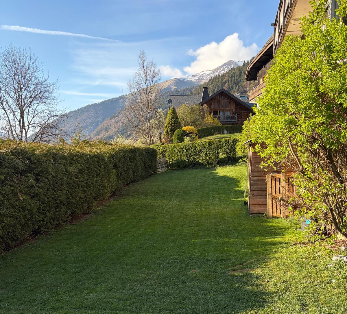 Neatly manicured garden with a lush green lawn bordered by a tall trimmed hedge, a wooden shed, and Alpine chalets and snow-capped mountains in the background under a blue sky.
